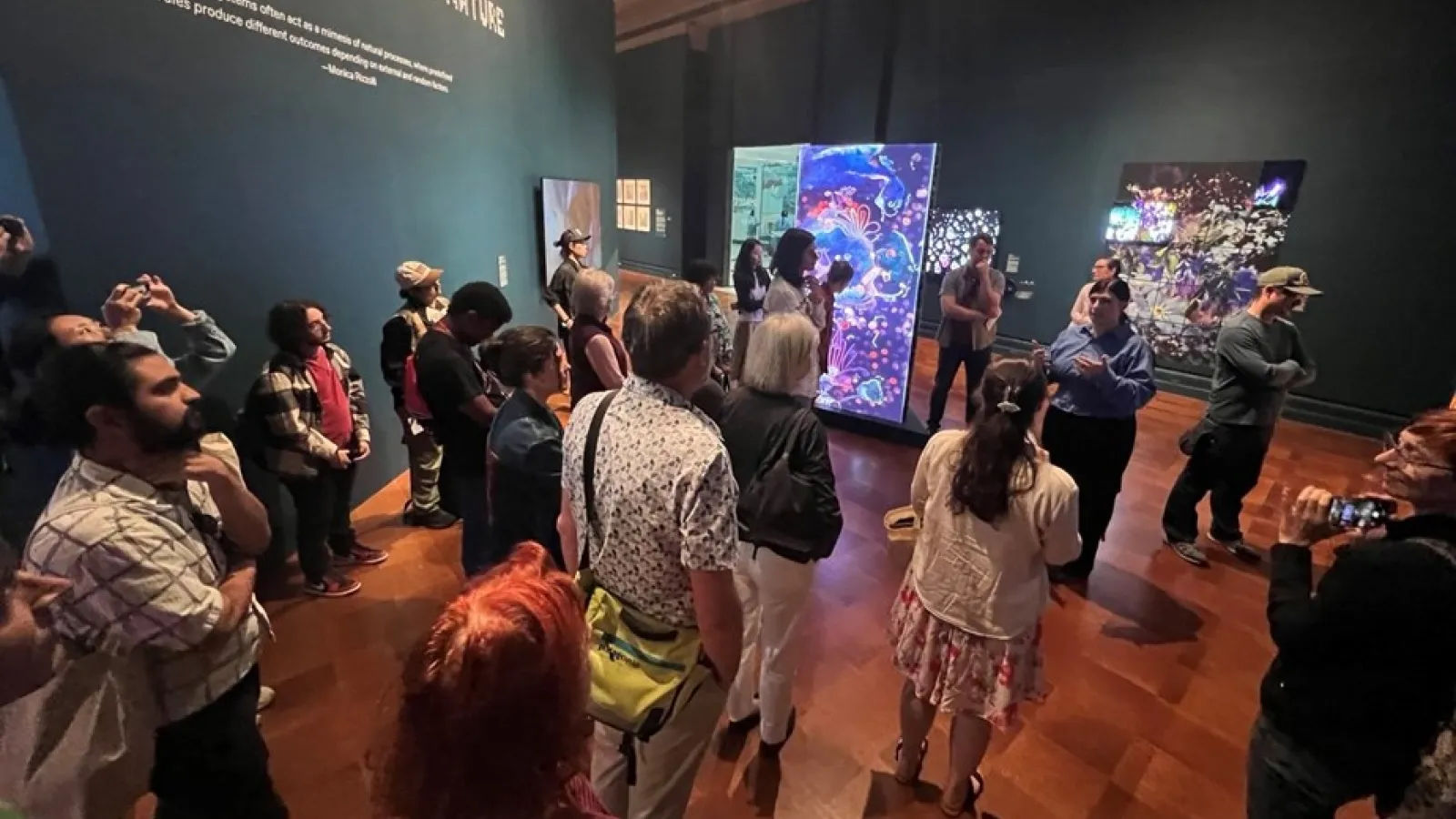 A group of approximately twenty people stand in a museum gallery with dark teal walls. They are attentively listening to a curator in a blue shirt who is speaking and gesturing. Behind the group, wall text reads "CODED NATURE," and several large, bright digital art screens display colorful, abstract floral imagery.