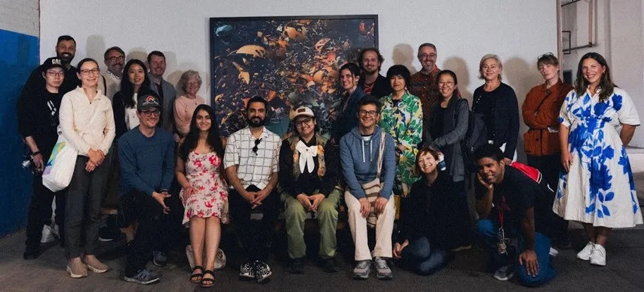 A group photograph of approximately twenty students and faculty members standing and sitting in two rows inside a gallery space. They are smiling and posing in front of a large, complex abstract digital artwork by Charles Csuri featuring fragmented shapes in dark, orange, and yellow tones.