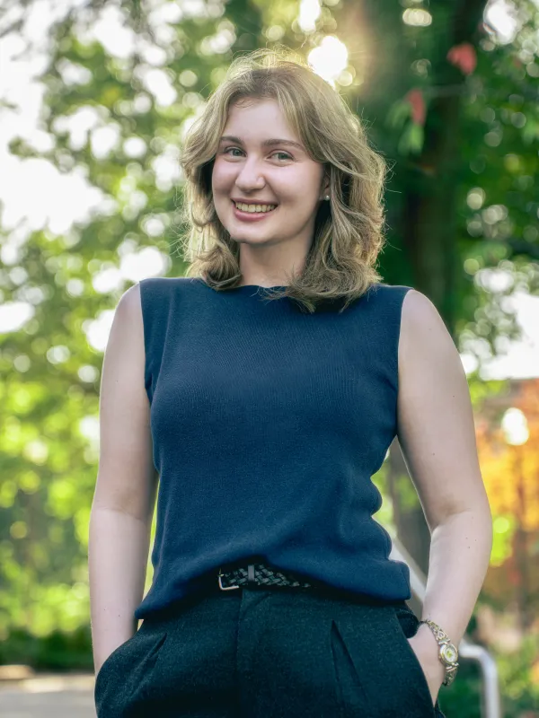 A young blonde woman stands with her hands in her pockets. She is smiling and weating a navy blue shirt.